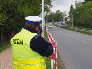 Policjant ruchu drogowego w białej czapce oraz żółtej kamizelce mierzy prędkość pojazdów za pomocą laserowego miernika prędkości.