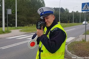 Zdjęcie kolorowe. Na zdjęciu policjant ruchu drogowego w kamizelce odblaskowej mierzy prędkość kierujących radarem laserowym. W dłoni trzyma także tarczę sygnalizacyjną. W tle widoczne jest przejście dla pieszych i nadjeżdżający samochód.