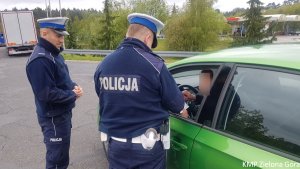 Zdjęcie kolorowe. Policjanci ruchu drogowego stoją przy samochodzie skoda Rapid. Jeden z policjantów wręcza kierującemu mandat karny.