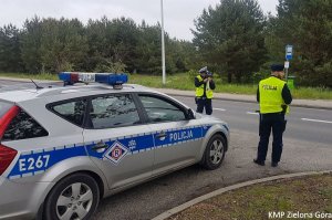 Zdjęcie kolorowe. Radiowóz ruchu drogowego zaparkowany przy drodze. W tle policjant ruchu drogowego mierzący prędkość. Obok stoi policjant z Samodzielnego Pododdziału Prewencji Policji.