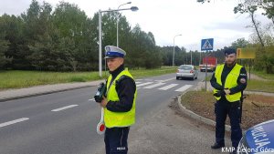 Zdjęcie kolorowe. Policjant ruchu drogowego stoi z laserowym miernikiem i przygotowuje się do mierzenia prędkości. Obok stoi policjant z Samodzielnego Pododdziału Prewencji Policji. W tle przejście dla pieszych i przejeżdżające samochody