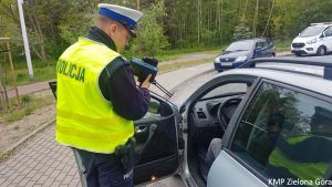 Zdjęcie kolorowe. Policjant ruchu drogowego stoi przy samochodzie marki seat i pokazuje kierującemu z jaką prędkością jechał.