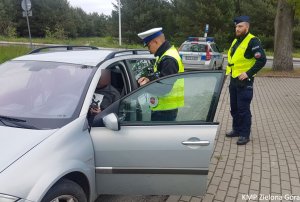 Zdjęcie kolorowe. Policjant ruchu drogowego i policjant z Samodzielnego Pododdziału Prewencji Policji stoją przy kontrolowanym samochodzie. W tle stoi radiowóz oznakowany.