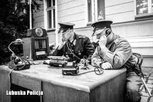 Czarno białe zdjęcie dwóch policjantów siedzących przy biurku, w tym jeden trzymający telefon stacjonarny