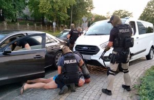 Dwóch zamaskowanych policjantów zakuwających w kajdanki mężczyznę leżącego na ziemi. Trzeci policjant wchodzi do auta w celu przeszukania