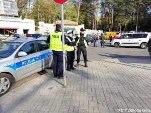 Policjanci stoją przy radiowozie i pilnują bezpieczeństwa na terenie parkingu cmentarza