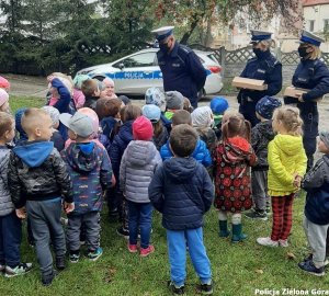 Policjanci ruchu drogowego rozmawiają z dziećmi, w tle policyjny radiowóz