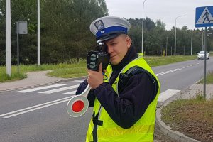 policjant w białej czapce i żółtej kamizelce odblaskowej stoi na drodze, w ręku trzyma laserowy miernik prędkości i tarczę do zatrzymywania kierujących, z tyłu za jego plecami widoczny znak drogowy oznaczający przejście dla pieszych i widoczny jadący biały samochód