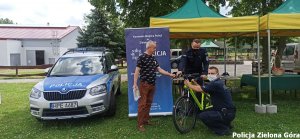 Dwóch funkcjonariuszy trzyma zielony rower, obok nich stoi starszy mężczyzna. W tle stoi zielony stragan i radiowóz policyjny.