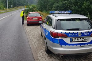 Przed zaparkowanym na poboczu radiowozem policji stoi w odległości 4 metrów czerwony samochód osobowy marki Peugeot.