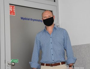 Na tle drzwi z napisem &quot;Wydział Kryminalny&quot; stoi mężczyzna w kraciastej, niebieskiej koszuli i czarnej maseczce.