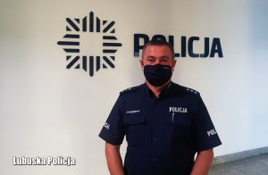 Funkcjonariusz policji w granatowej koszulce i maseczce stoi na tle białej ściany z granatowym napisem &quot;POLICJA&quot;