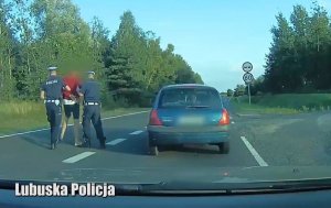 Dwóch funkcjonariuszy policji trzymający kierowcę granatowego Renault ubranego w czerwoną koszulkę.