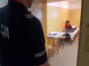 Funkcjonariusz policji stojący przy otwartym pomieszczeniu dla osób zatrzymanych, w którym na białym łóżku siedzi skulona kobieta.