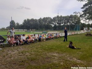 Pan funkcjonariusz rozmawia z dziećmi na temat bezpieczeństwa w czasie wakacji na tle boiska piłkarskiego. Obok funkcjonariusza leży jego pies służbowy.