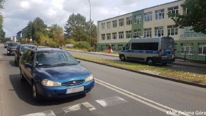 Na pierwszym planie niebieski samochód osobowy, za nim trzy inne samochody. Na drugim planie policyjny bus transporter stoi przed budynkiem szkoły