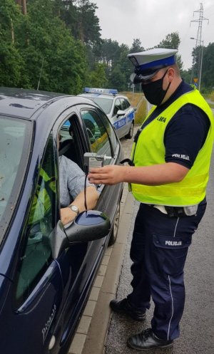 policjant w kamizelce odblaskowej i białej czapce sprawdza trzeźwość kierującego w granatowym samochodzie przy użyciu urządzenia alcoblow