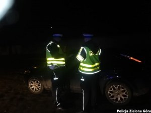 w nocy dwaj policjanci w kamizelkach odblaskowych i w białych czapkach kontrolują trzeźwość kierującego