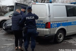 Policyjny radiowóz typu bus. Przy tylnych drzwiach dwoje policjantów oraz zatrzymany mężczyzna.