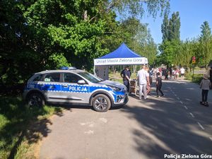 Radiowóz policyjny a tle stragan policyjny wraz z dwójką policjantów.