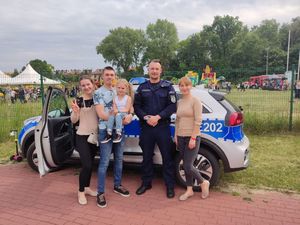 Mężczyzna trzymający dziewczynkę na rękach wraz dwoma kobietami stoją obok umundurowanego Policjanta, w tle radiowóz.