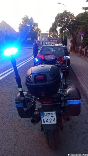 Stojący na sygnałach błyskowych motor, obok Policjant Wydziału Ruchu Drogowego podejmujący czynności służbowe wobec kierowcy samochodu osobowego.