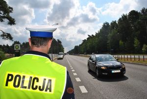 Policjant drogówki, jezdnia, samochód w ruchu, samochody na poboczu, słoneczny dzień, znak stacji paliw.