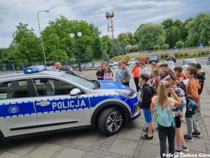 Samochód policyjny, Policjant obok samochodu, grupka dzieci, słoneczny dzień, plac przed Komendą Miejską.