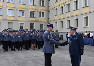 Dwóch policjantów ściska sobie dłonie na tle grupy policjantów stojących na baczność.