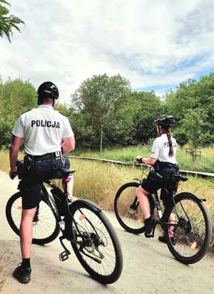 Policjant i policjantka ubrani w białe t-shirty i szorty z napisem POLICJA pełnią służbę na rowerach.