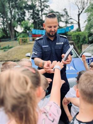 Policjant rozdaje dzieciom opaski odblaskowe w kolorze srebrno-szarym.