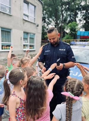 Policjant rozdaje dzieciom opaski odblaskowe w kolorze srebrno-szarym.