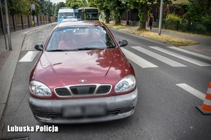 Samochód w kolorze ciemnym czerwonym stoi na jezdni, przy przejściu dla pieszych. Za ciemnoczerwonym pojazdem stoi policyjny radiowóz.