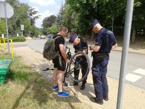 Dwóch policjantów w umundurowaniu z napisem POLICJA sprawdzają legalność pochodzenia jednośladu należącego do stojącego obok mężczyzny.