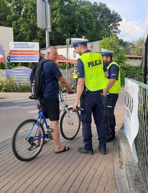 Dwóch policjantów w mundurach i kamizelkach z napisem POLICJA rozmawiają z rowerzystą.