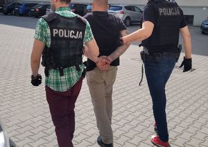 Dwóch policjantów w kamizelkach z napisem POLICJA prowadzi zatrzymanego mężczyznę. Zatrzymany mężczyzna ubrany jest w czarną koszulkę oraz spodnie w kolorze cielistym.