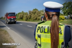 Policjantka w umundurowaniu z napisem POLICJA mierzy, z jaką prędkością nadjeżdża pojazd ciężarowy w kolorze czerwono-czarnym.