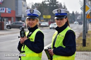 Dwie policjantki w umundurowaniu z napisem POLICJA stoją przy jezdni. Jedna z policjantek trzyma w ręce radar mierzący prędkość nadjeżdżających pojazdów.