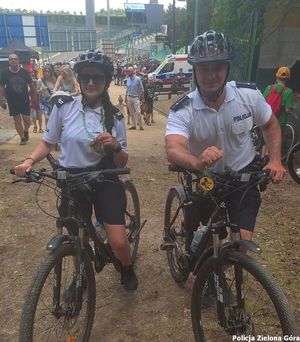 Policjant i policjantka pełnią służbę na rowerach. Ubrani są w biały t-shirt z napisem POLICJA, oraz czarne szorty.