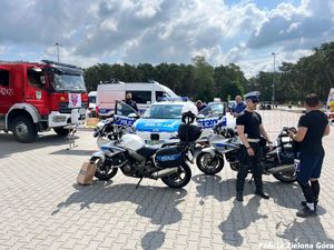 Policjant stoją przy motorach policyjnych rozmawiając z mężczyzną. W tle widoczny jest radiowóz policyjny, przy nim stoi policjant w mundurze z napisem POLICJA.