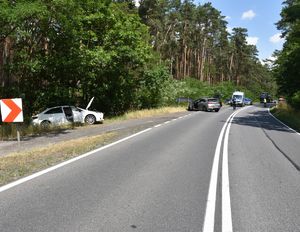 Fotografia ukazująca kolizję drogową na drodze krajowej. W tle widoczny jest radiowóz policyjny oraz funkcjonariusze Policji.