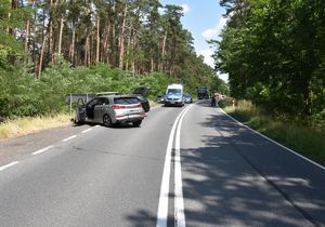 Kolizja drogowa między srebrnym, a czarnym samochodem. W tle widoczny jest radiowóz policyjny.