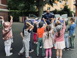 Policjant w mundurze z napisem POLICJA stoi z grupą dzieci. W tle widoczny jest radiowóz policyjny.