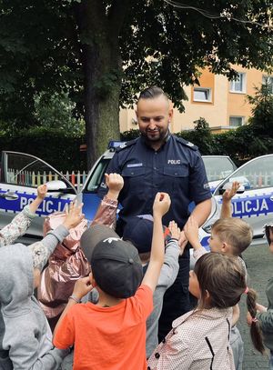 Policjant stoi z grupą dzieci. Dzieci ochoczo podnoszą ręce w kierunku funkcjonariusza.