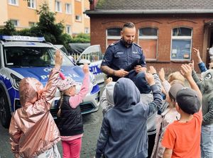 Policjant w mundurze z napisem POLICJA rozdaje grupie dzieci opaski odblaskowe. Policjant stoi na tle radiowozu policyjnego.