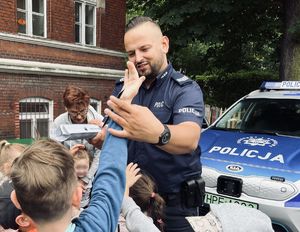 Policjant w mundurze z napisem POLICJA zakłada chłopcu opaskę odblaskową. Policjant stoi na tle radiowozu policyjnego.