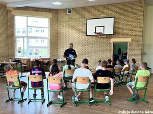 Policjant prowadzi zajęcia z grupą dzieci. W ręce trzyma ulotki.