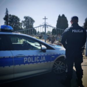 Policjant stoi przy radiowozie obok cmetarza.