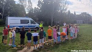 Dzieci stoją na przeciwko policjantki za którą znajduje się radiowóz policyjny.