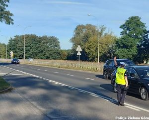 Policjant podchodzi do samochodu osobowego z ręką w górze zwróconą w stronę nadjeżdżającego samochodu.